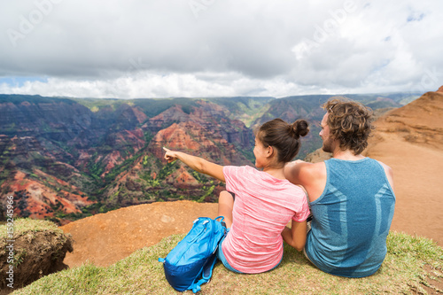 People hiking in Hawaii hikers pointing at Kauai. happy hiker couple healthy lifestyle outdoors looking at Waimea canyon view. Young couple resting sitting in nature in Kauai, Hawaii, USA.