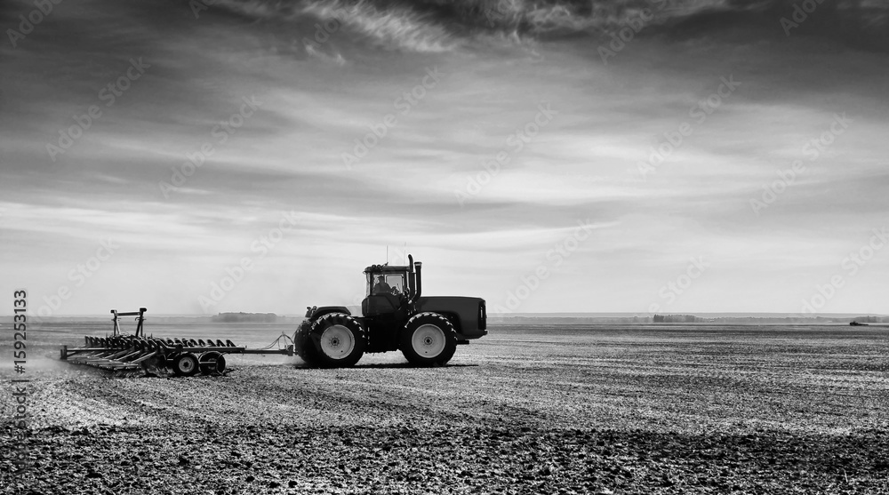 Fototapeta premium A tractor with attached cultivator working the land in a rural summer black and white countryside landscape