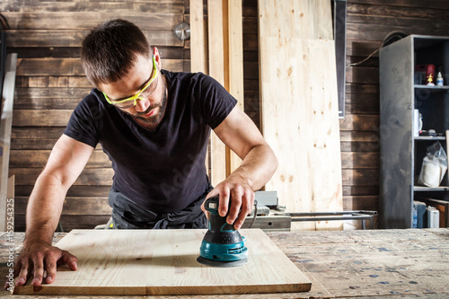 Foto Young man builder carpenter equals polishes wooden board with a  random orbit sa