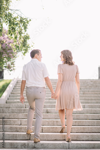 beautiful couple on a walk, the girl in the beige dress and the guy wearing light clothing go up the stairs back, holding hands, Park, summer, nature photo
