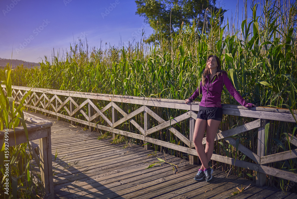 Mujer joven apoyada en la barandilla de una pasarela de madera en la naturaleza con ropa deportiva Mujer joven apoyada en la barandilla de una pasarela de madera en la naturaleza con ropa deportiva