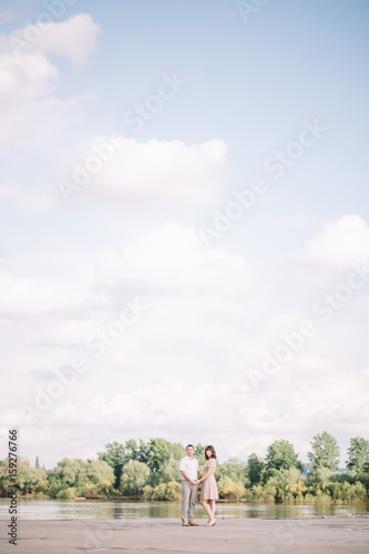 love story, beautiful couple a man and a woman in bright clothes, dress walking along the river Bank,are holding hands, vertical, blue sky with white clouds, summer, nature