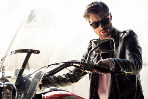 Handsome stylish young man in sunglasses and leather jacket sitting on motorcycle and looking at camera