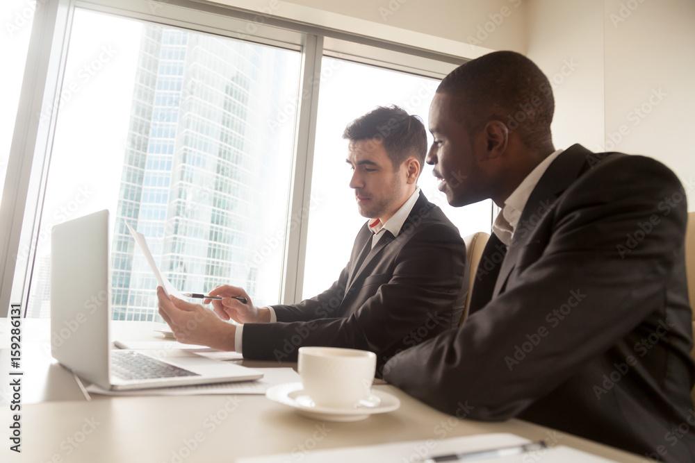 Fototapeta premium Side view of two multiracial business partners in formal wear discussing document on meeting sitting at desk, black and white businessmen studying contract terms, explaining idea, planning project