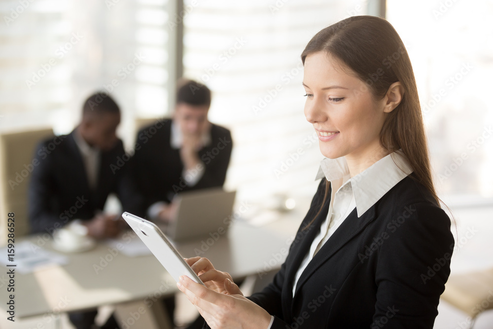 Young attractive businesswoman holding tablet, looking at screen, standing in office with colleagues at background, organizing scheduling meeting, using business app for managing time and daily tasks Young attractive businesswoman holding tablet, looking at screen, standing in office with colleagues at background, organizing scheduling meeting, using business app for managing time and daily tasks