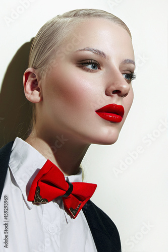 Beautiful young showy girl in a man s style. Smoothly combed blond hair  bright makeup - red lipstick  dark shadows  smooth shiny skin. Clothes - a black jacket  a white shirt  a red bow tie. Fashion