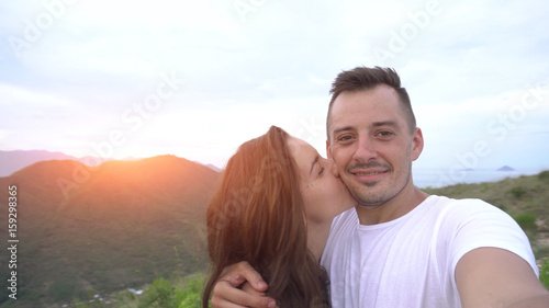 Smiling couple taking selfie in mountains, aerial city view. Woman kisses man.