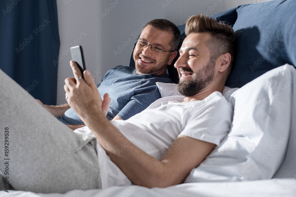 Fototapeta premium Happy gay couple using smartphone while lying together on bed
