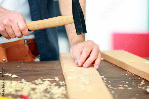 Carpenter driving nail into wooden board in workshop, closeup