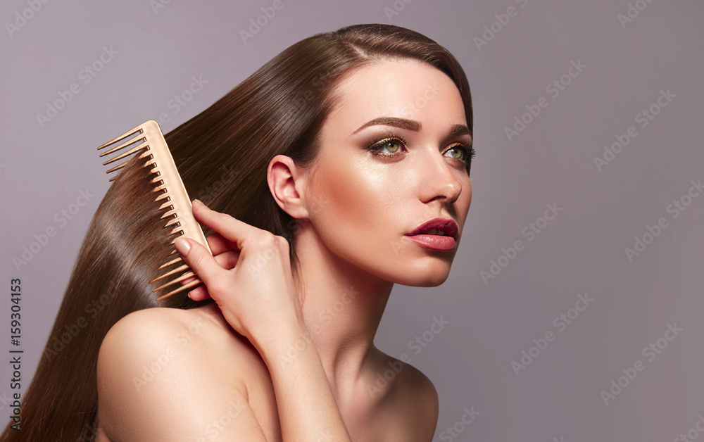 Fototapeta premium A beautiful young girl with long shiny silky hair sits and combs her hair with a golden comb. Cosmetics for hair, care, spa, beauty, fashion. Beauty saloon. Smooth skin. Advertising. Radiance, shine.