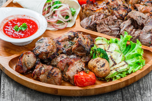 Assorted delicious grilled meat and vegetables with fresh salad and bbq sauce on cutting board on wooden background close up. Hot Meat Dishes. Top view
