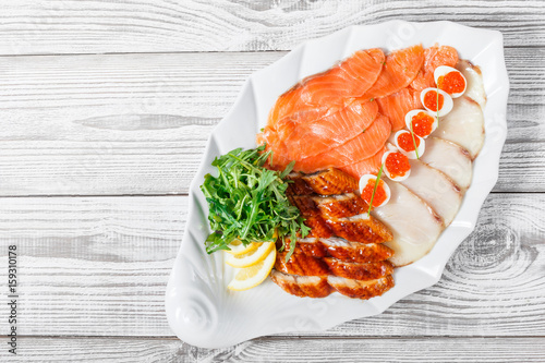 Seafood platter with salmon slice, smoke sturgeon, quail eggs with red caviar, slices fish fillet, decorated with arugula and lemon on wooden background close up. Mediterranean appetizers. Top view