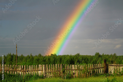 In the summer after rain in the village behind an old wooden fence in a blue sky a bright rainbow. Nature is erased, the colors have become bright!