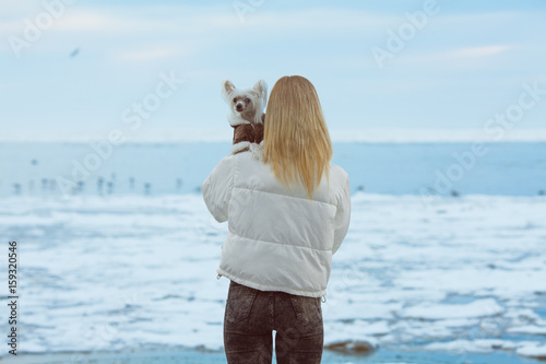 Winter vacation concept. Couple of friends standing on beach with flying seagulls near water. Girl hugs her Chinese crested dog. Hipster style. Happy together. Outdoor shot