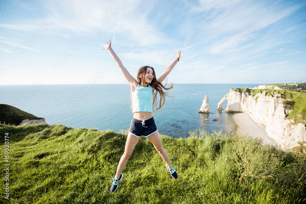 Young playful woman in sportswear jumping outdors on the beautiful rocky coastline background near Etretat town in France Young playful woman in sportswear jumping outdors on the beautiful rocky coastline background near Etretat town in France
