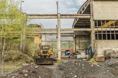 Outside view of old abandoned factory ruin with broken windows and glass rusty steel structure with yellow bulldozer.