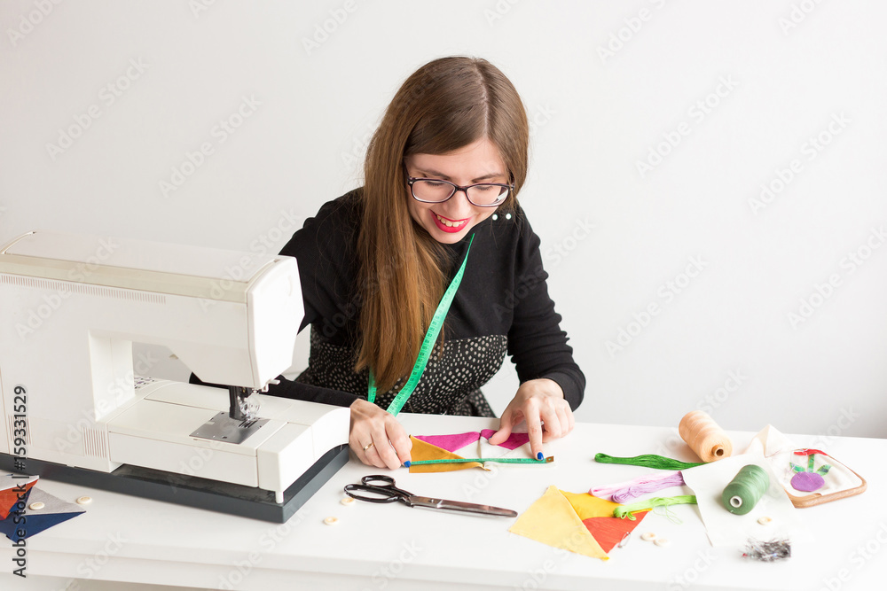 needlework and quilting in the workshop of a young woman, a tailor on white background - smiling female tailor measuring fabric with tape measure and preparing for sewing machine needlework and quilting in the workshop of a young woman, a tailor on white background - smiling female tailor measuring fabric with tape measure and preparing for sewing machine