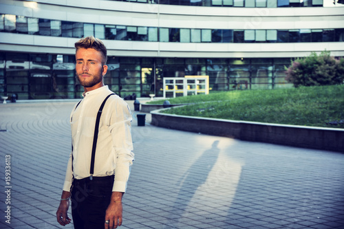 Blond Young Man Next to Modern Building in City, Wearing White Shirt and Suspenders, Standing