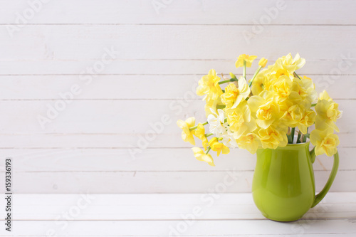 Bright yellow spring daffodils or narcissus flowers in pitcher on light wooden background.
