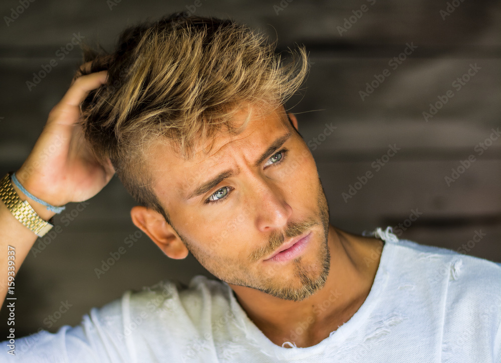 Obraz premium Handsome blond, blue eyed young man leaning against wood planks wall outdoor and looking away to a side
