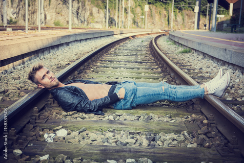 Confident stylish young man with hands in pockets lying on train rails. photo