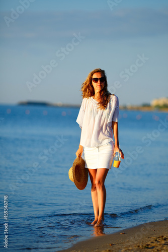 Young woman with orange juice in hand in white clothes on the beach