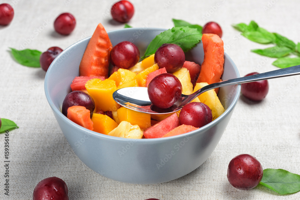 Mixed fruit salad in the blue bowl. Healthy sliced fresh fruits on a linen grey background. Red berries, green leaves and spoon on table