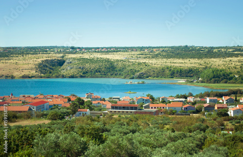 Crkva Sveti Duh church in Posedarje in Novigradsko More bay in blue azure water against the background of a hill, green trees and houses with red roofs. Warm cloudless day in Croatia