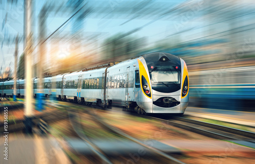 High speed train in motion at the railway station at sunset. Modern european intercity train on the railway platform with motion blur effect. Industrial scene with moving passenger train on railroad