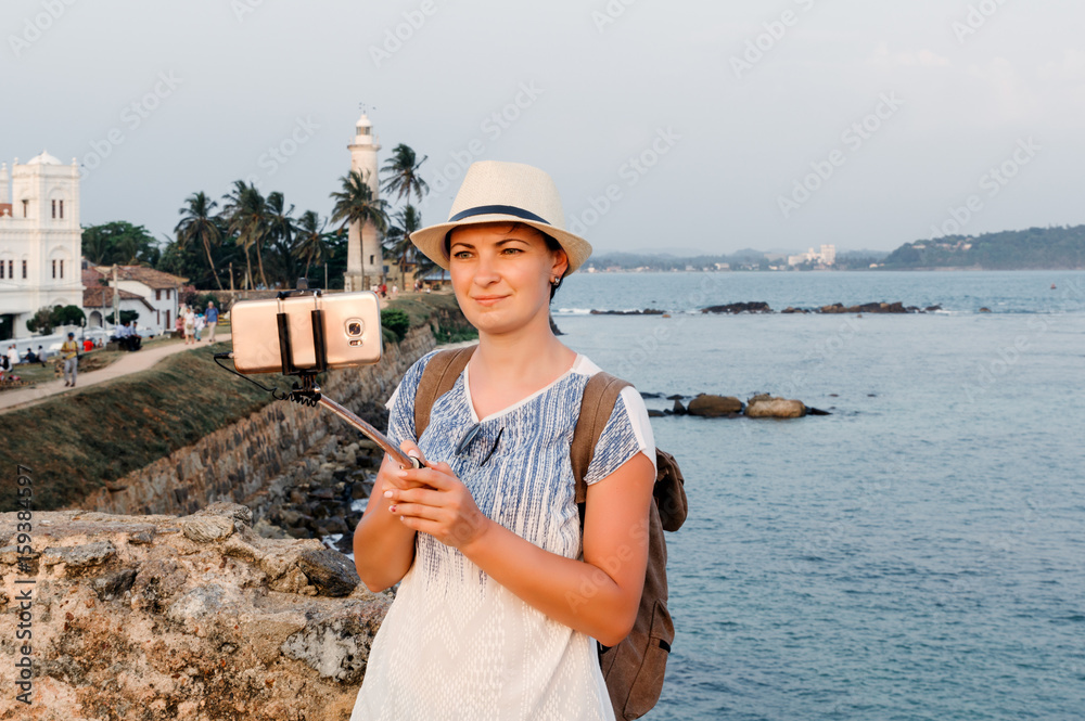Girl traveler with backpack doing selfie on mobile phone. Light house at Galle Dutch Fort 17th centurys ruined dutch castle. That is unesco listed as a world heritage site in Sri Lanka.
