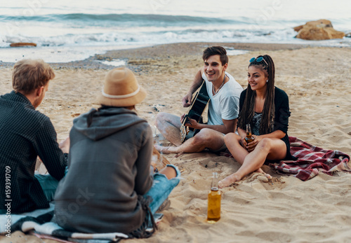 group of young and cheerful friends sitting on beach and lit bonfire. One man is playing guitar. Music on Wild beach