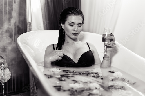 Side view of Sexy brunette woman relaxing in a hot bath with flowers and champagne in hand. she is wearing black sexual lingerie. woman in bath. Her eyes are closed. black and white photo. bw