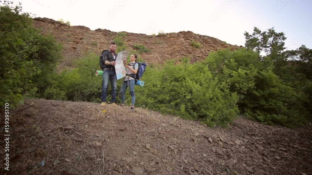 Two tourists with backpacks looking for a road using the map standing on top of a mountain. Young tourist couple traveling on holidays in mountain looking at map in search of attractions. Two tourists with backpacks looking for a road using the map standing on top of a mountain. Young tourist couple traveling on holidays in mountain looking at map in search of attractions.