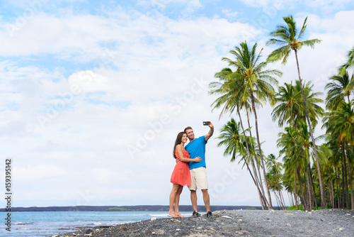 Selfie couple on Hawaii beach vacation with palm trees and volcanic black sand in Big island of Hawaii, USA. Hawaiian holidays getaway. Happy people on summer holidays.