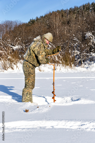Fisher drills a hole in the ice. Winter fishing on the background of a frozen lake.