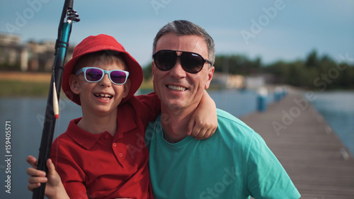 Cute happy young boy wearing trendy sunglasses posing arm in arm with his father at a lake in front of a jetty holding a large fishing rod. © Framestock