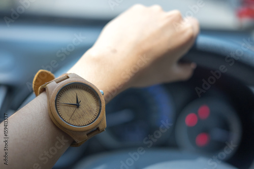 business woman hand wearing wooden wrist watch. her holding steering wheel car for driving and have car interface status are background. 