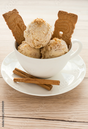 Three scoops of homemade Gingerbread Ice Cream in a white porcelain cup decorated with Spekulatius and cinnamon sticks on light brown wooden background. Backlit.