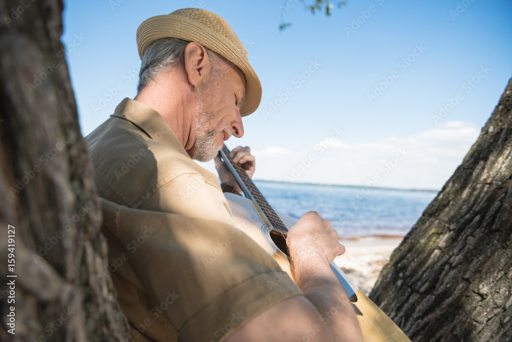 Senior man in hat leaning on tree and playing guitar at riverside