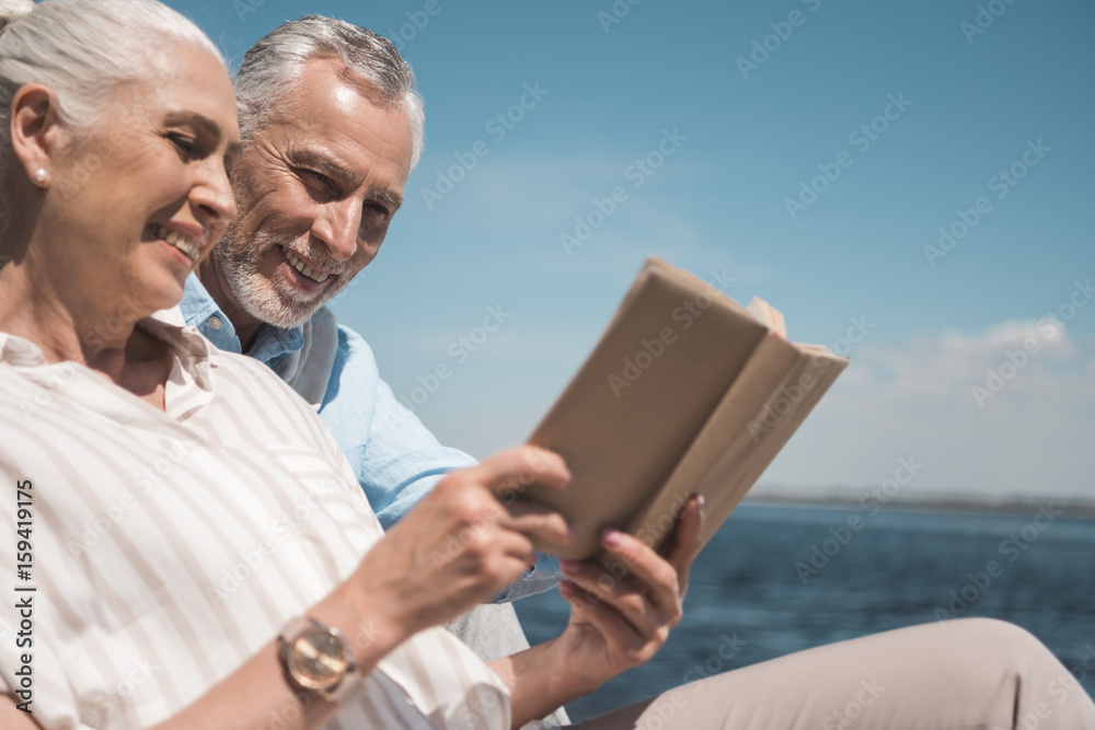 smiling elderly couple reading book on the quay at daytime