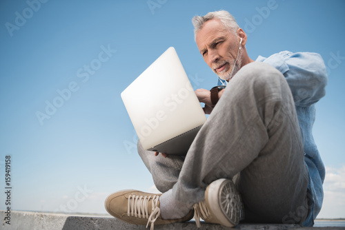 pensive elderly man working on laptop and listening music in earpods outdoors