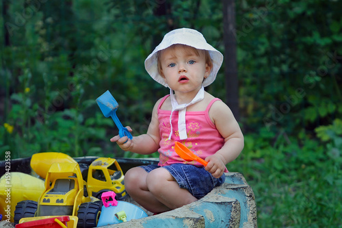 A small charming girl is playing in the sandbox in the backyard of her house. Infant is sitting with a plastic spatula among toys in a sandbox made of a large wheel from a tractor.