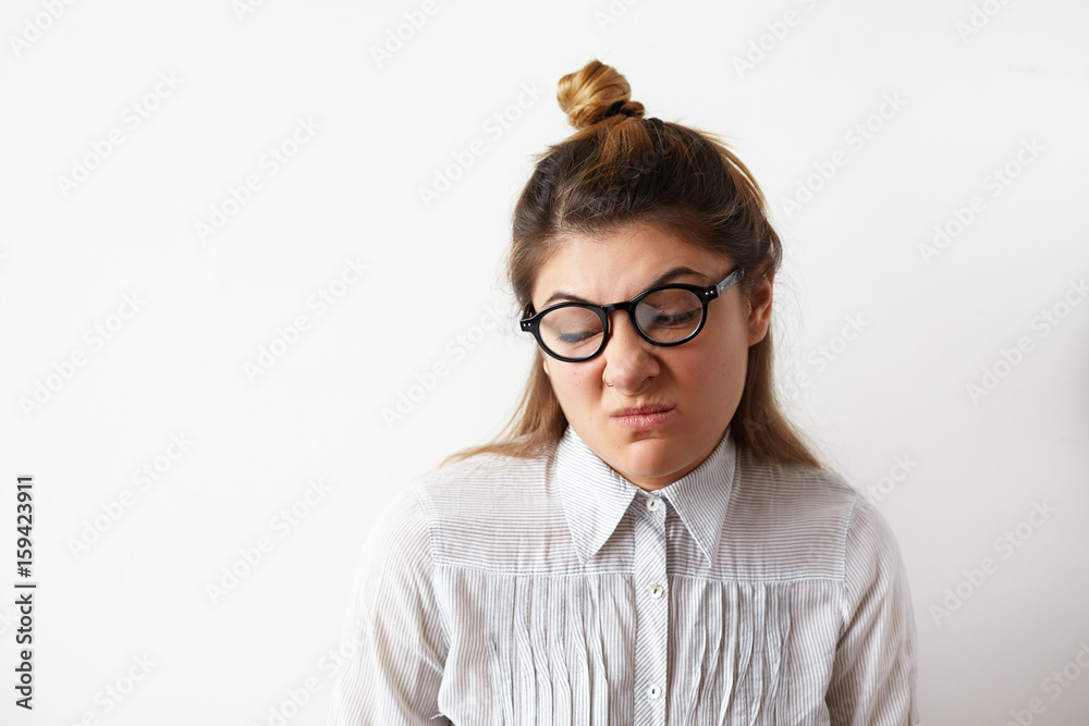 Headshot of young businesswoman female with hair knot wearing shirt and glasses looking aside with suspicious and disgusted expression on her face. Caricature of negative human emotions, and feelings
