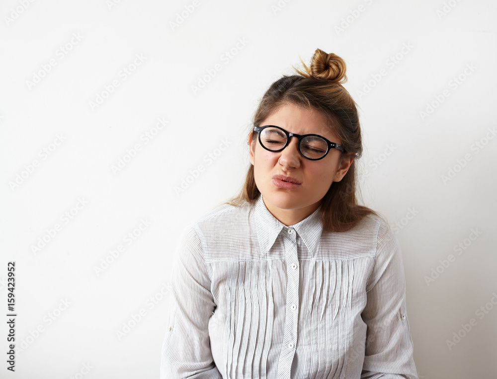 Portrait of stressful young woman with hair bun in formal clothes and eyeglasses blinked for a headache or migraine. Tired woman having some problems looking stressed isolated on white wall background Portrait of stressful young woman with hair bun in formal clothes and eyeglasses blinked for a headache or migraine. Tired woman having some problems looking stressed isolated on white wall background