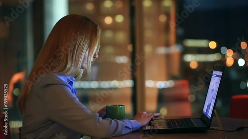 Beautiful blonde business woman working overtime at night in executive office. City lights are visible in background from a large window