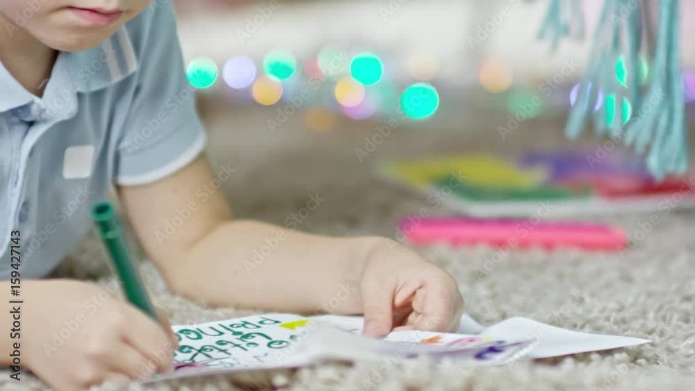 Tracking shot of little boy lying on the floor and writing wishes in greeting card with felt tip pen Tracking shot of little boy lying on the floor and writing wishes in greeting card with felt tip pen