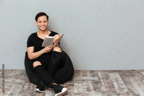 Happy young asian man sitting isolated over grey wall
