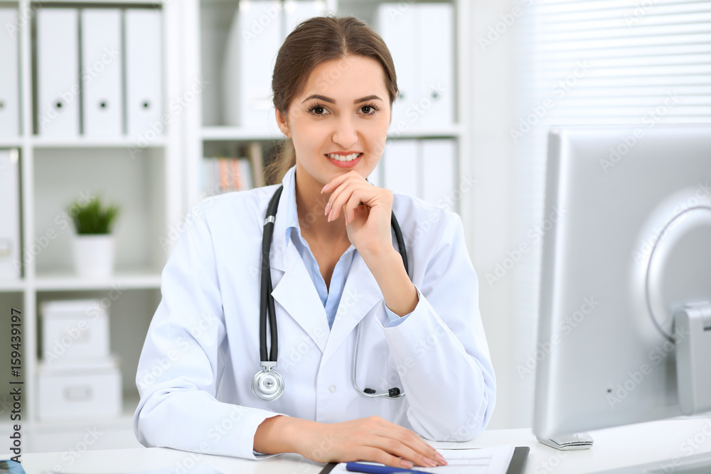 Latin american female doctor sitting at the table and working by computer at hospital office. The physician or therapist makes a diagnosis. Health care, medicine and patient service concept Latin american female doctor sitting at the table and working by computer at hospital office. The physician or therapist makes a diagnosis. Health care, medicine and patient service concept