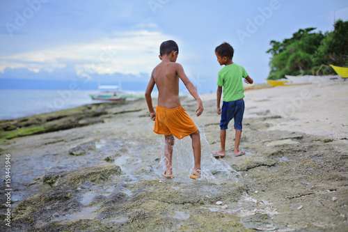 two little asian boys splashing their feet in shallow water ahead of oncoming waves of the sea at sunset, Island, Philippines