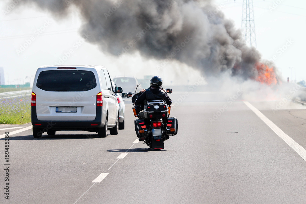 Car crash / accident / road disaster concept. Vehicles passing a car burning with a thick black smoke on the highway